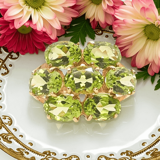 a gold ring with a cluster of oval cut green gemstones on a gold and white plate with pink flowers behind it