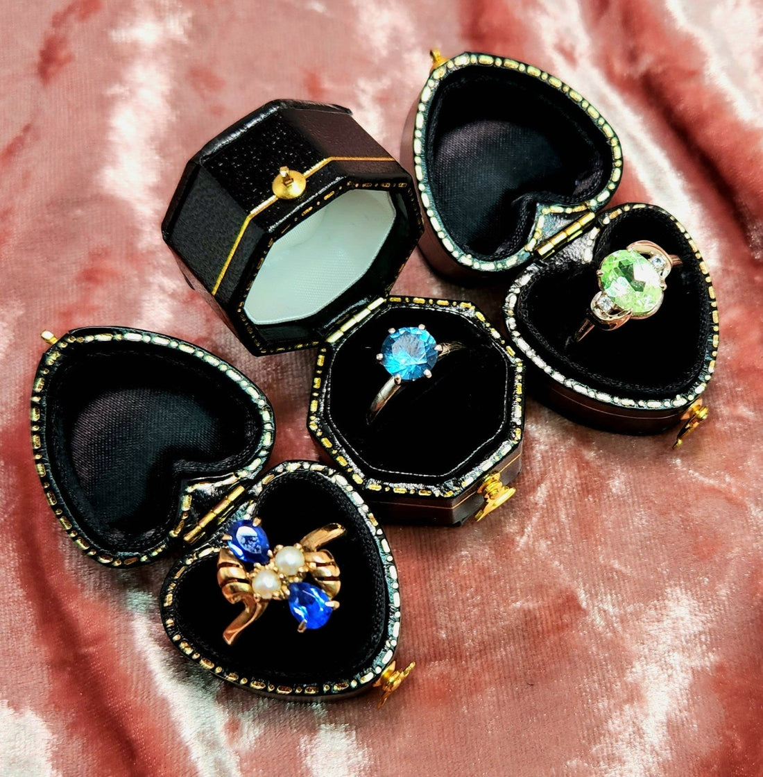 three blue and green rings in black heart jewelry boxes on pink velvet