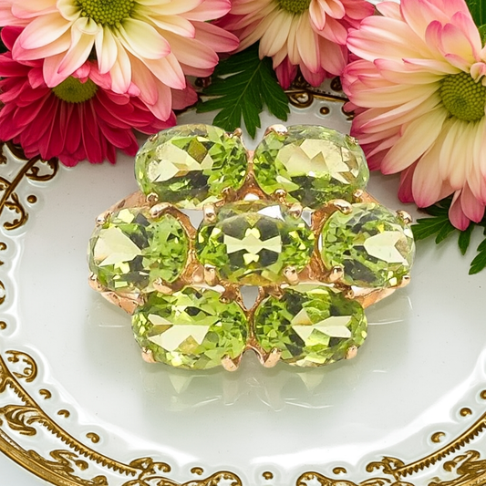 a gold ring with a cluster of oval cut green gemstones on a gold and white plate with pink flowers behind it