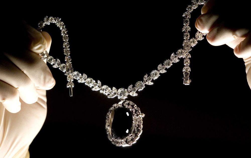 gloved hands holding up a large diamond necklace with a large black diamond