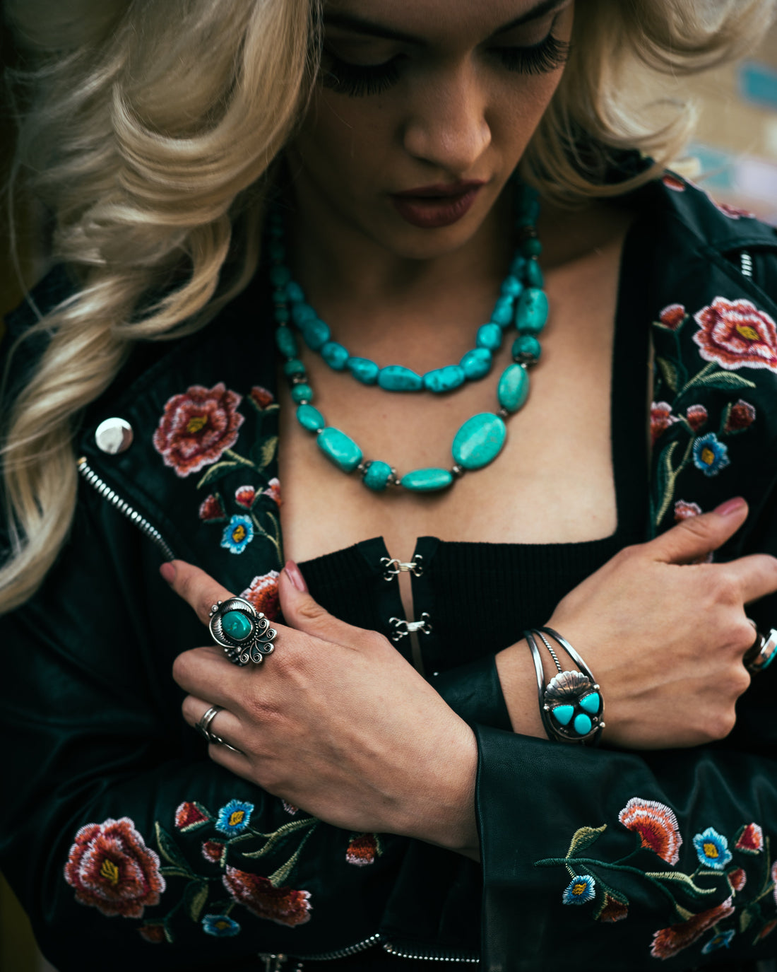 a blonde woman with a black coat covered in pink roses and wearing a lot of turquoise jewelry