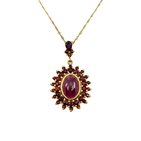 A gold pendant necklace featuring a central oval ruby cabochon surrounded by a sunburst of smaller round red gemstones, suspended from a delicate gold chain on a plain white background.